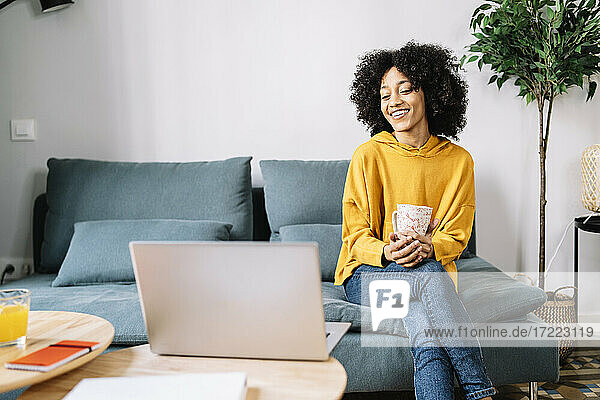Lächelnde Frau mit Tasse  die auf einen Laptop schaut  während sie zu Hause sitzt
