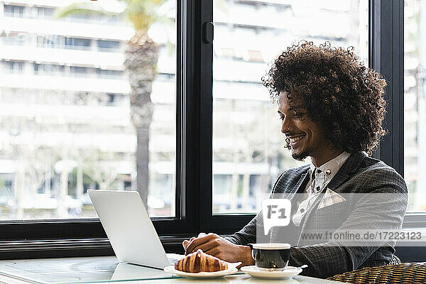 Lächelnder männlicher Unternehmer mit Laptop neben Croissant und Kaffeetasse in einem Café