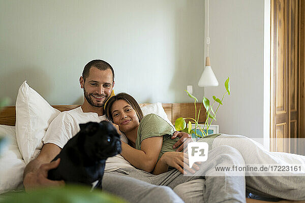 Happy couple with Pug dog lying on bed at home