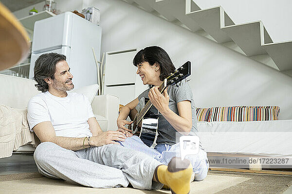 Lächelnde Frau  die mit einem Mann Gitarre spielt  während sie zu Hause sitzt