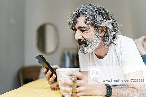 Mann  der ein Mobiltelefon benutzt und einen Kaffeebecher hält  während er zu Hause liegt