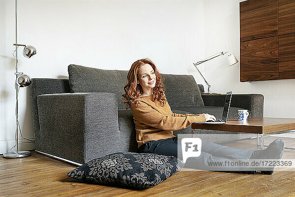 Frau in voller Länge mit Laptop sitzend und wegschauend im Wohnzimmer zu Hause