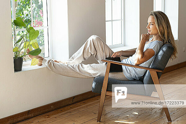 Mittlere erwachsene Frau mit Hand am Kinn träumt  während sie am Fenster im Wohnzimmer sitzt