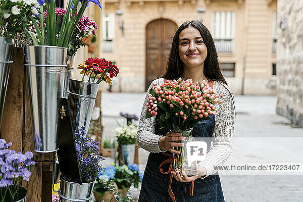 Junge Blumenhändlerin hält eine Vase mit Hypericum-Blumen  während sie in einem Blumenladen steht