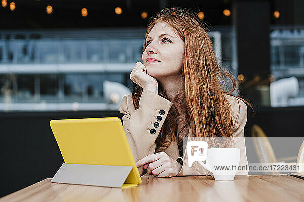 Schöne Frau mit Hand am Kinn träumt  während sie vor einem digitalen Tablet und einer Kaffeetasse im Café sitzt