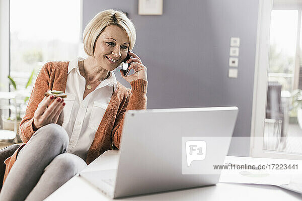 Lächelnde Unternehmerin  die mit ihrem Smartphone telefoniert und etwas isst  während sie zu Hause arbeitet