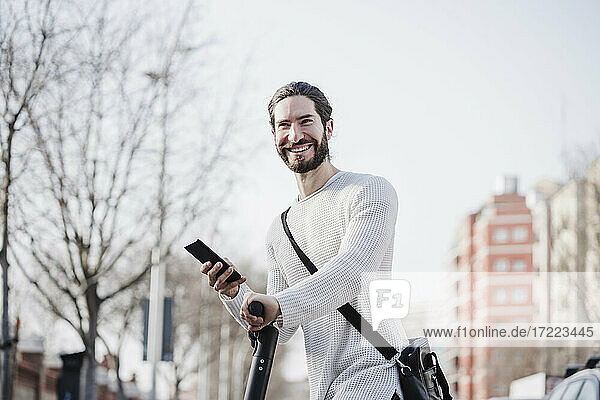 Glücklicher männlicher Hipster  der sein Smartphone auf einem Motorroller hält