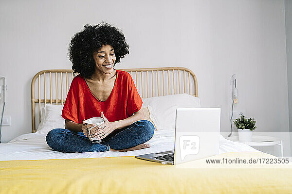 Junge Frau mit Tasse  die auf einen Laptop schaut  während sie zu Hause auf dem Bett sitzt