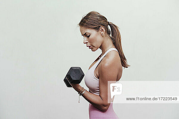 Female sportsperson lifting dumbbell while exercising by white wall