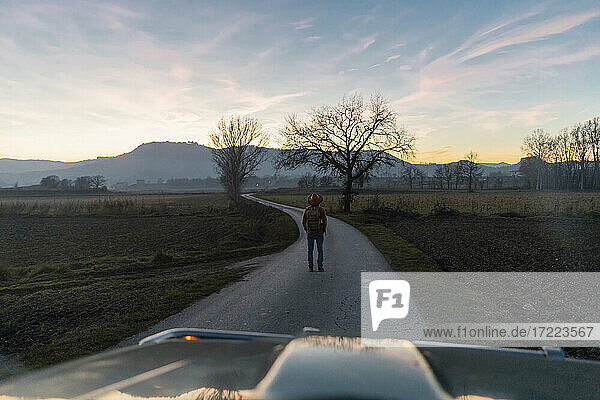 Mann steht bei Sonnenuntergang auf einer Landstraße vor einem Auto