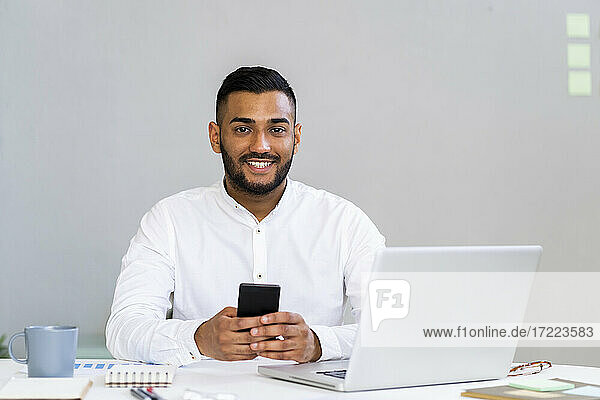 Lächelnder junger männlicher Unternehmer mit Smartphone am Laptop  der am Schreibtisch im Büro sitzt