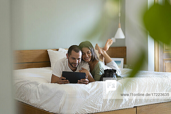 Lächelndes junges Paar mit Mops-Hund  das ein digitales Tablet benutzt  während es zu Hause auf dem Bett liegt