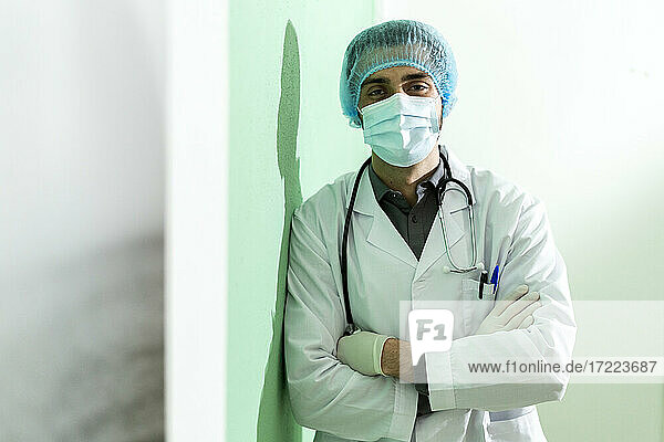 Männlicher Arzt mit Chirurgenmütze  der mit verschränkten Armen im Krankenhaus steht  während COVID-19
