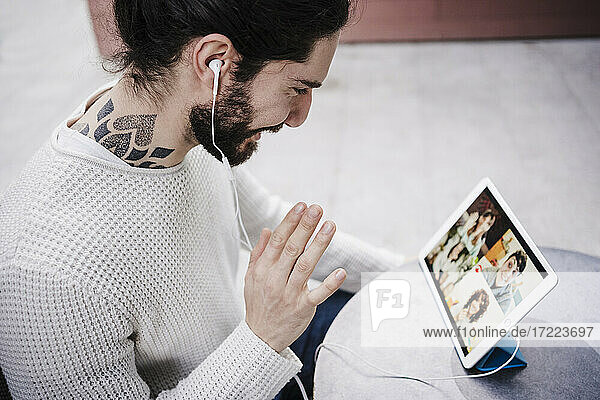 Mann mit Tätowierung winkt mit der Hand während eines Videoanrufs über ein digitales Tablet in einem Straßencafé
