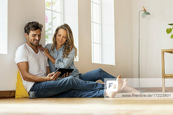 Lächelndes Paar  das ein digitales Tablet benutzt  während es zu Hause auf dem Boden sitzt