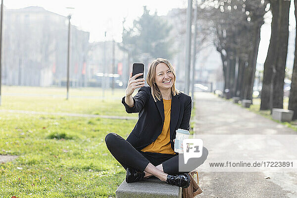 Fröhliche Geschäftsfrau  die ein Selfie mit ihrem Mobiltelefon macht  während sie auf einer Bank im Park sitzt