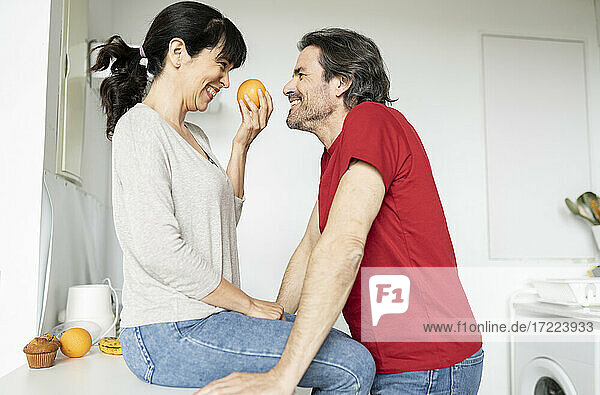 Woman holding orange looking at man while sitting on kitchen counter at home