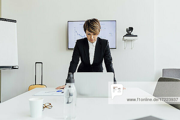 Ernste Geschäftsfrau  die auf einen Laptop schaut  während sie sich auf dem Schreibtisch gegen einen Projektionsschirm im Büro lehnt