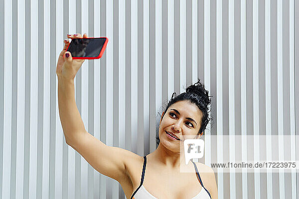 Lächelnde Sportlerin  die ein Selfie mit ihrem Handy vor einer gestreiften Wand macht