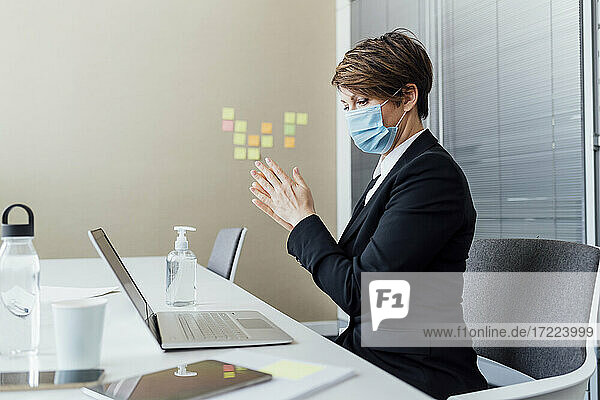 Weibliche Fachkraft mit Gesichtsschutzmaske reibt sich die Hände  während sie ein Desinfektionsmittel am Schreibtisch im Büro benutzt