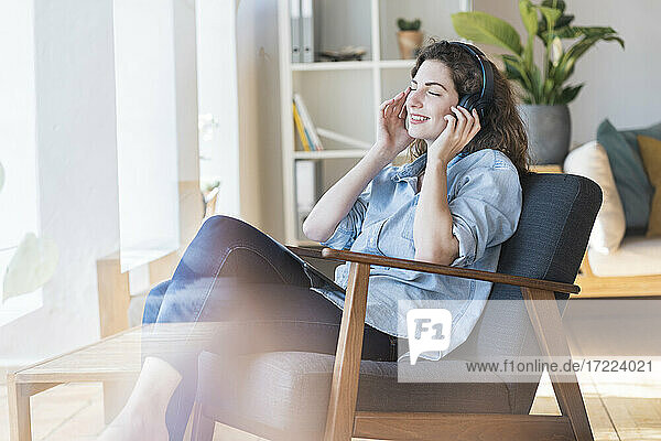 Lächelnde schöne Frau hört Musik über Kopfhörer  während sie auf einem Stuhl zu Hause sitzt