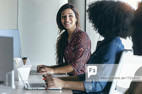 Lächelnde weibliche Fachkraft  die einen Kollegen bei der Arbeit am Laptop im Büro ansieht