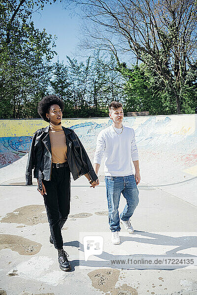 Multi ethnic couple walking while holding hands at skateboard park during sunny day