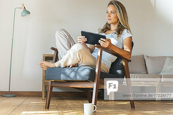 Contemplating woman holding digital tablet while sitting on armchair in living room