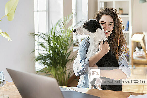 Happy businesswoman cuddling dog at desk in home office