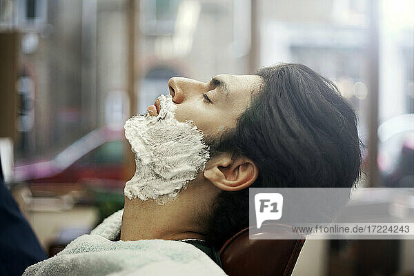 Profile of male customer sitting in barber shop with shaving cream on face