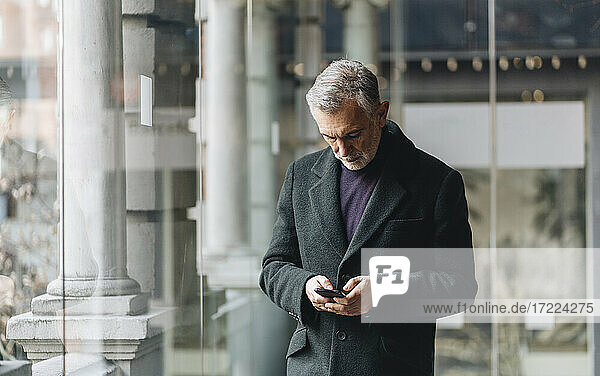 Reifer  modischer Mann  der ein Mobiltelefon an einer Glaswand benutzt