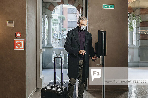 Mann mit Gesichtsschutzmaske  der Handdesinfektionsmittel aufträgt  während er neben seinem Gepäck im Hotel steht