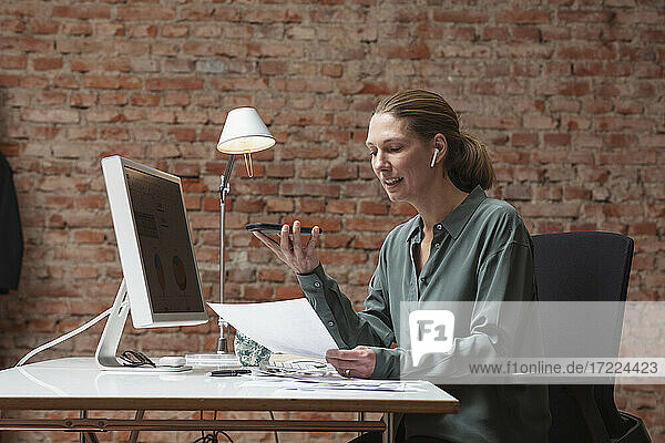Berufstätige Frau spricht über drahtlose Kopfhörer  während sie ein Dokument am Schreibtisch hält