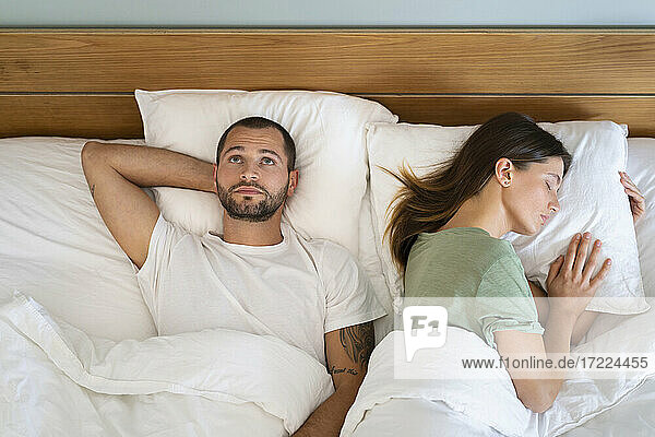 Junge Frau schläft neben einem besorgten Mann  der an die Decke starrt  während er zu Hause auf dem Bett liegt