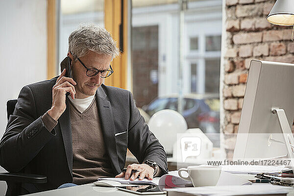 Reifer Geschäftsmann  der ein Dokument betrachtet  während er am Arbeitsplatz mit einem Mobiltelefon spricht