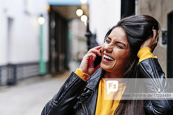 Cheerful young woman squinting while talking on mobile phone in city
