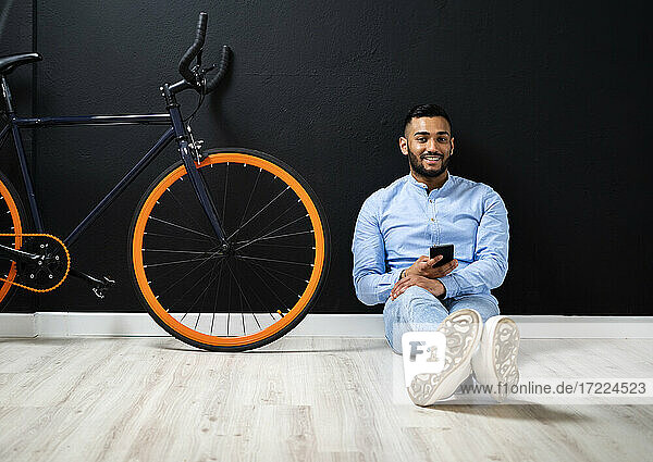Handsome man with smart phone sitting near bicycle by wall at studio