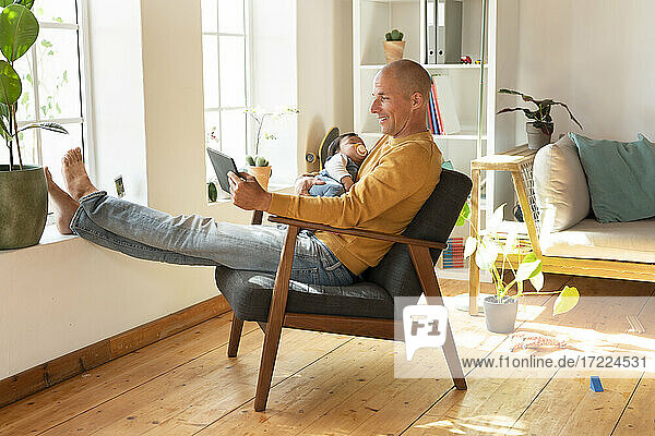 Lächelnder Vater  der ein digitales Tablet benutzt und sein Baby hält  während er zu Hause im Sessel sitzt