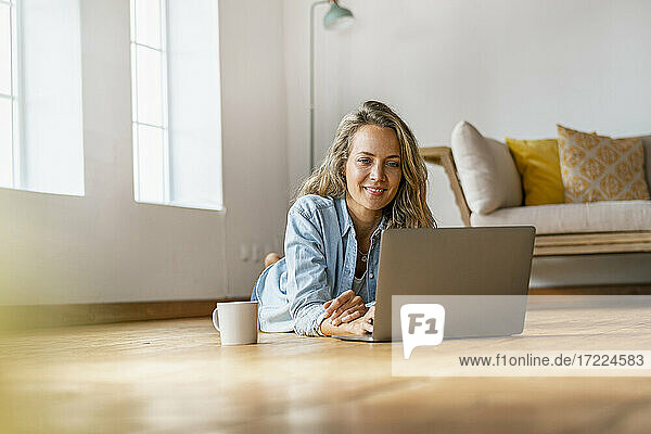 Lächelnde Frau auf Hartholzboden vor einem Laptop zu Hause liegend