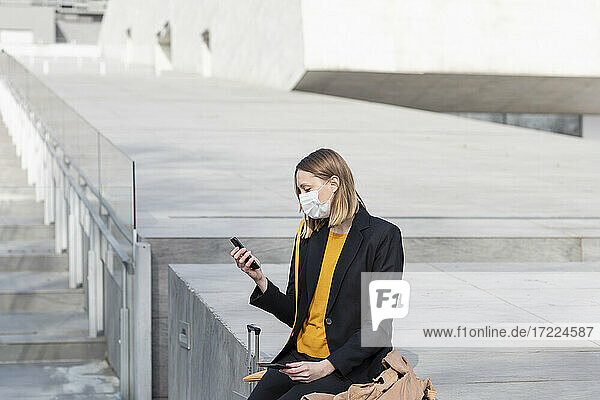 Female entrepreneur with face mask using mobile phone while sitting by staircase