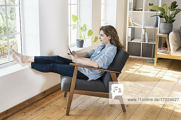 Entspannte junge Frau mit digitalem Tablet  die mit hochgelegten Füßen auf einem Stuhl am Fenster im Wohnzimmer sitzt