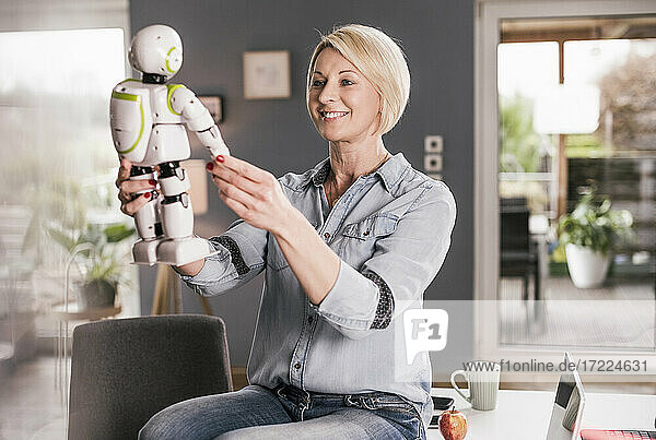 Lächelnde Geschäftsfrau  die ein Modell eines Roboters hält  während sie auf einem Tisch im Heimbüro sitzt