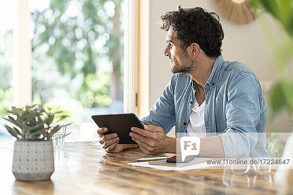 Male freelancer day dreaming while holding digital tablet at home office