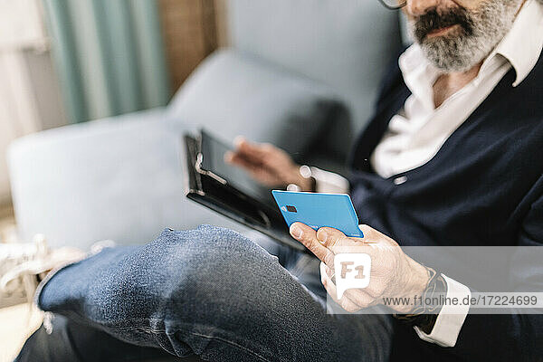 Ein älterer Mann hält eine Kreditkarte in der Hand  während er ein digitales Tablet zu Hause benutzt