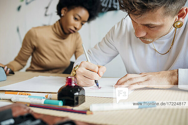 Hipster-Mann schreibt mit Füllfederhalter auf Papier  während er neben einer Frau im Atelier sitzt