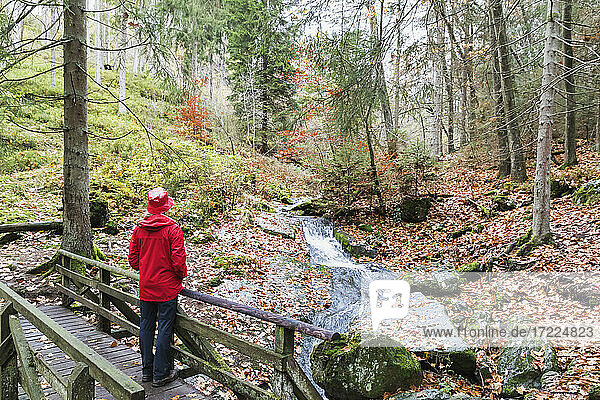 Ältere Frau mit rotem Mantel und Hut steht auf einer Brücke im Wald