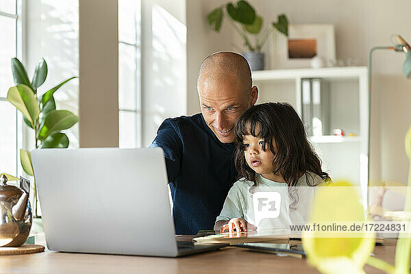 Vater arbeitet am Laptop  während er mit seinem Kleinkind im Home Office sitzt