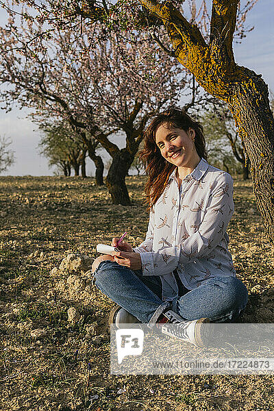 Lächelnde junge Frau sitzt mit Notizblock unter einem Mandelbaum