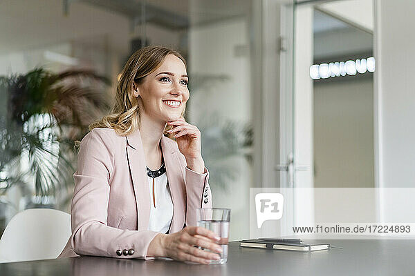 Lächelnde Geschäftsfrau schaut weg  während sie am Schreibtisch im Büro Wasser trinkt