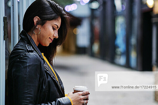 Lächelnde Frau mit Einweg-Kaffeebecher an der Wand lehnend